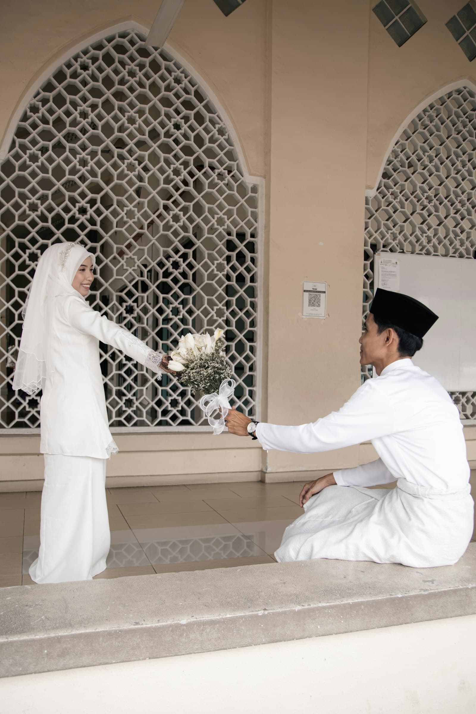 A couple in white at a mosque, the groom kneeling to offer a bouquet
