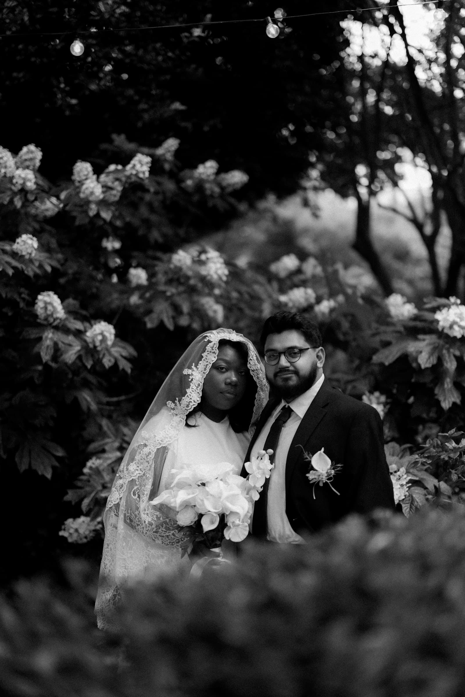 A black-and-white portrait of a bride in a veil and her groom among flowers