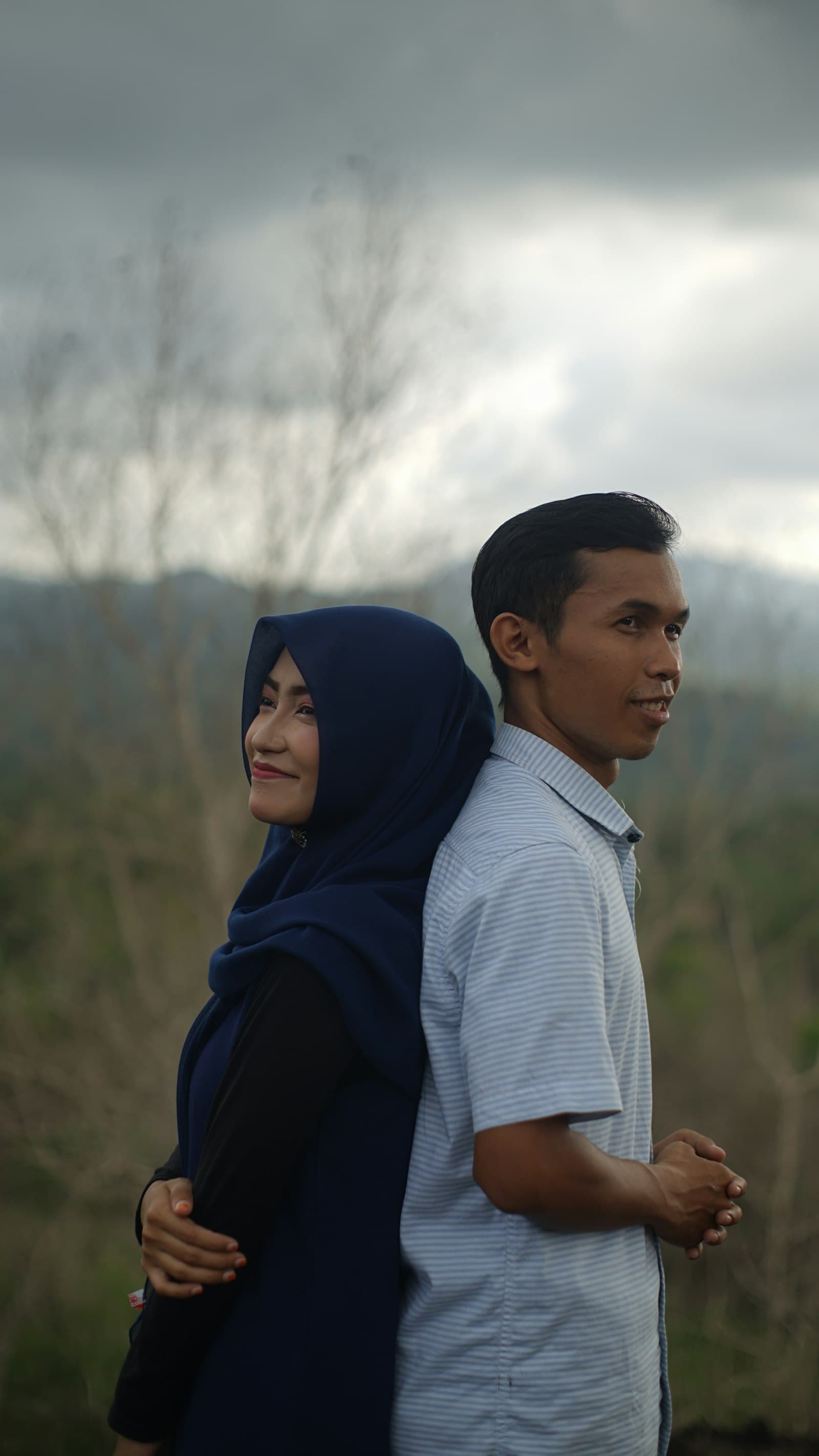 A couple standing back to back outdoors, the bride in a navy hijab